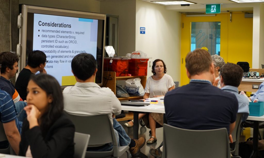 Image of a group of people around a table, discussing. A slide titled "Considerations" is projected on a screen. 